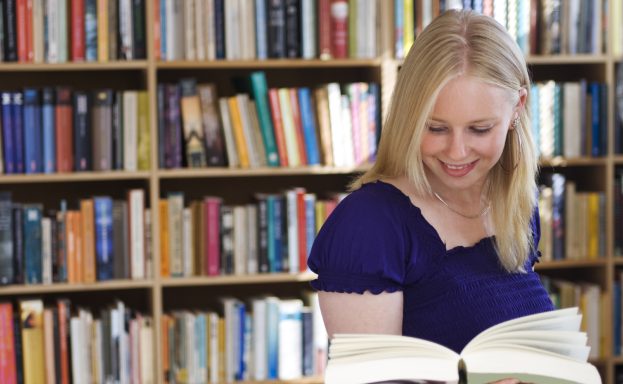 Lächelnde Frau liest ein Buch vor einem Bücherregal mit vielen bunten Büchern.