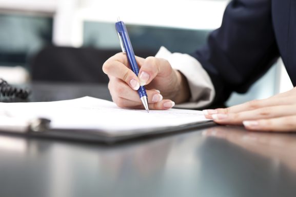 Eine Hand schreibt mit einem blauen Stift auf ein Blatt Papier in einem Ordner.