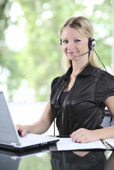 Junge Frau mit Headset am Laptop, lächelnd vor einem Fenster mit natürlichem Licht.