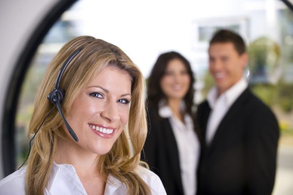Frau mit Headset lächelt, im Hintergrund zwei Personen in formeller Kleidung.