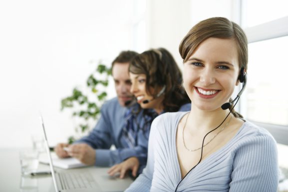 Lächelnde Frau mit Headset, zwei Personen im Hintergrund, alle arbeiten an Laptops.