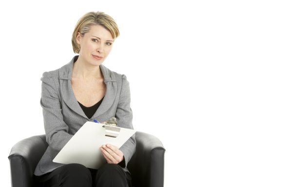 Frau im grauen Blazer sitzt auf einem Stuhl und hält eine Schreibunterlage.