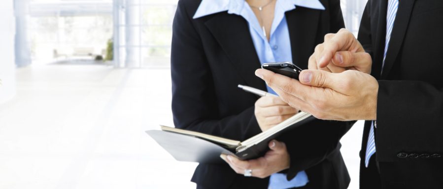 Zwei Personen in Anzügen, eine mit Smartphone, die andere mit Notizbuch.
