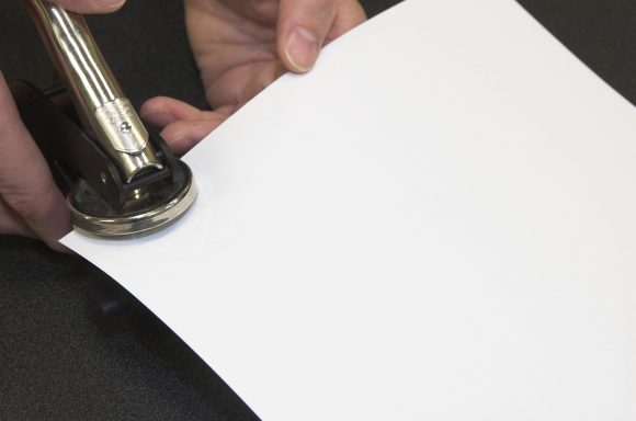 Eine Hand hebt einen Locher über ein Blatt Papier auf einer dunklen Oberfläche.