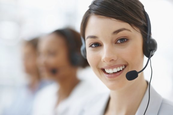 Lächelnde Frau mit Headset in einem Callcenter, im Hintergrund weitere Mitarbeiter.