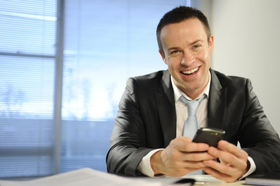Lächelnder Mann in Anzug hält ein Smartphone in der Hand und sitzt am Tisch.