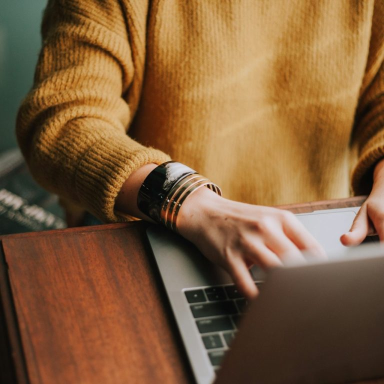 Person mit gelbem Pullover arbeitet an einem Laptop auf einem Holztisch.