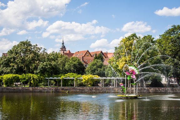 Park mit Teich, Bäumen und blühenden Pflanzen vor einem historischen Gebäude.
